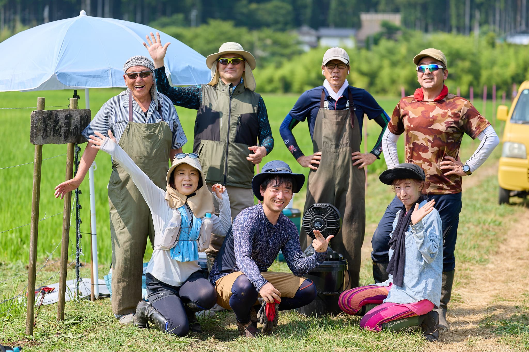 青田を背に、麦わら帽子や日除け姿で笑顔を見せる田んぼ仲間の集合写真