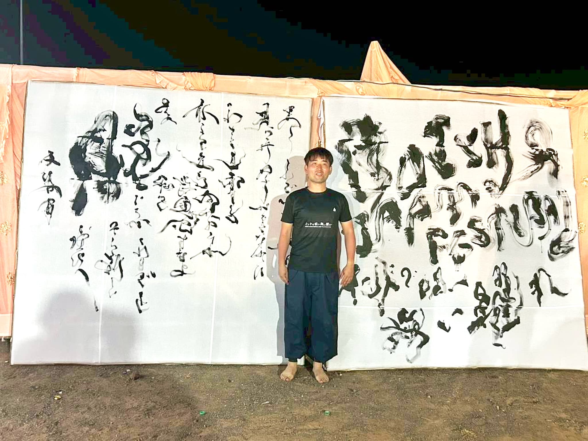 夜の屋外会場で、大型書作品2枚（左に「脱ぐ」、右に祭事の大字）の前に立つ作者