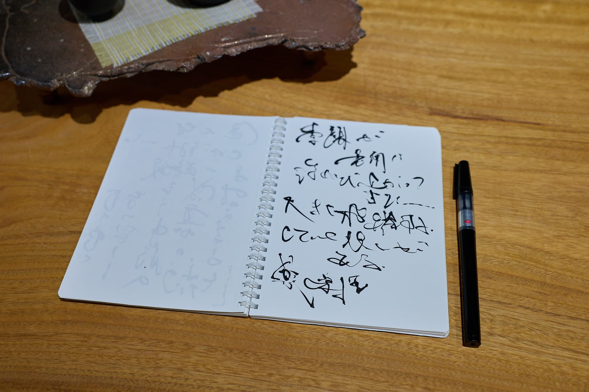 木のテーブルに開いたスケッチブックに、筆ペンで書きためた言葉の下書き