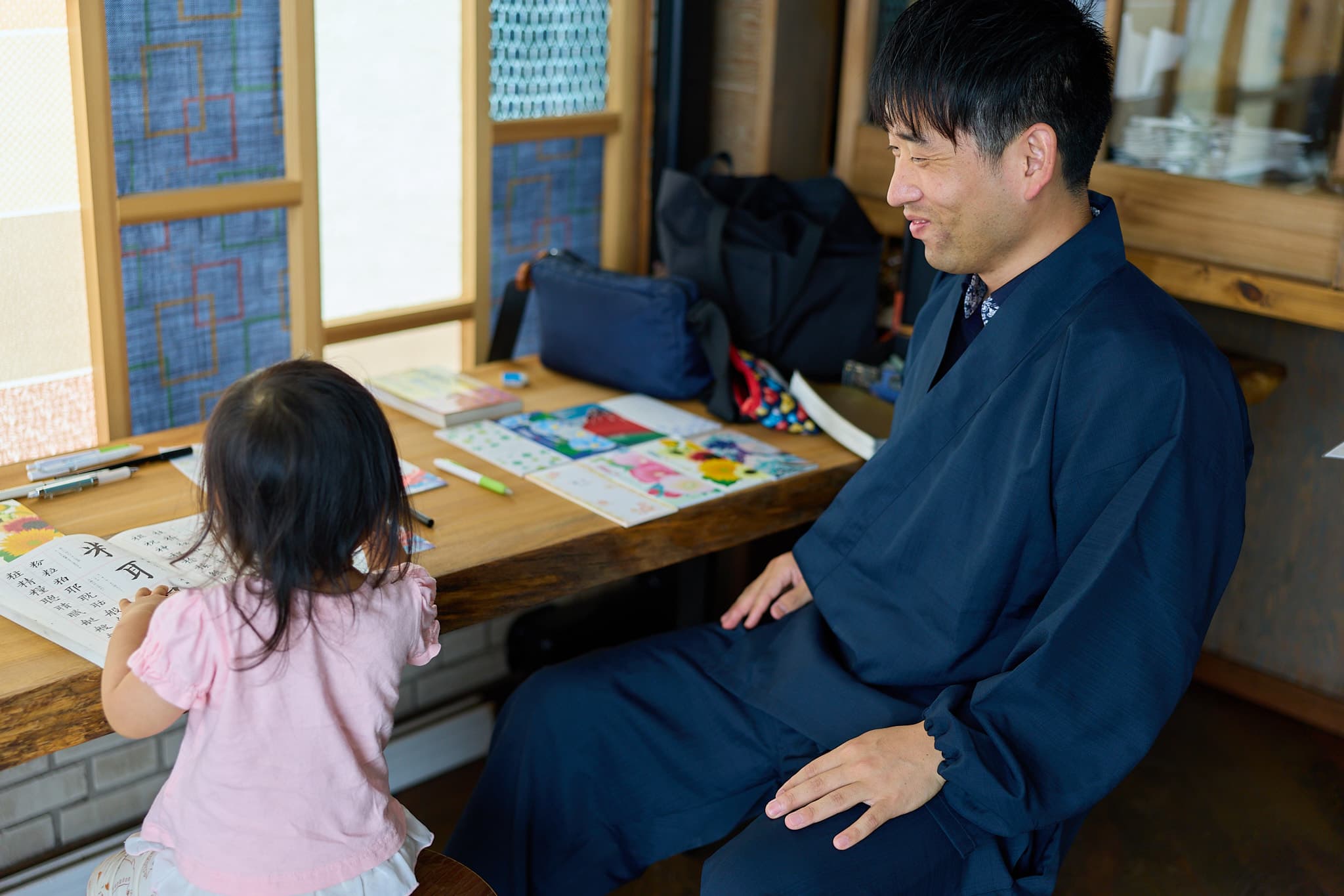 紺の着物姿の森下友喜が、習字のお手本を手にした小さな女の子と和やかに向き合う様子