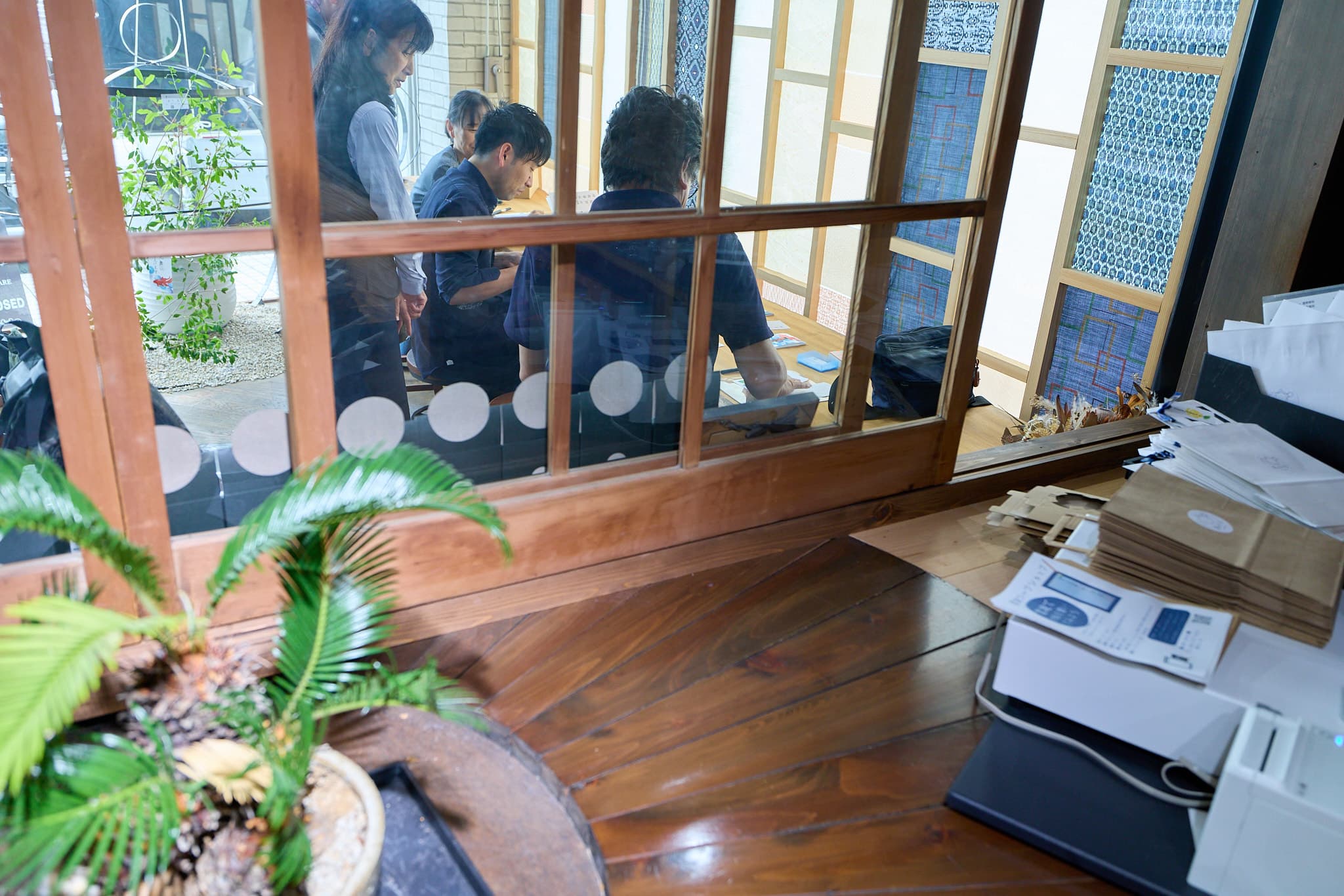 和室のギャラリーで、窓越しに来場者が集うイベント会場の風景。ソテツの鉢と什器が並ぶ