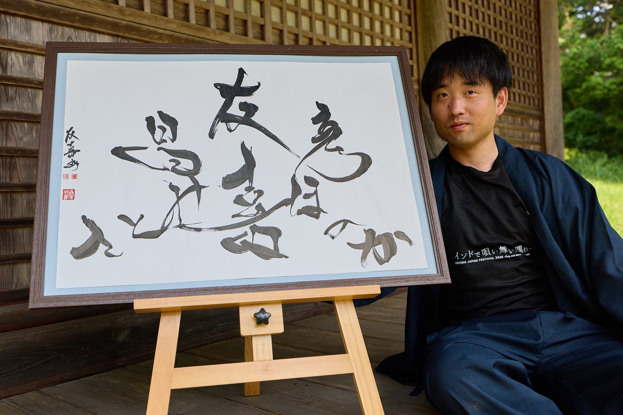 和歌を揮毫した横長の額装作品と、その傍らに座る作務衣姿の森下友喜