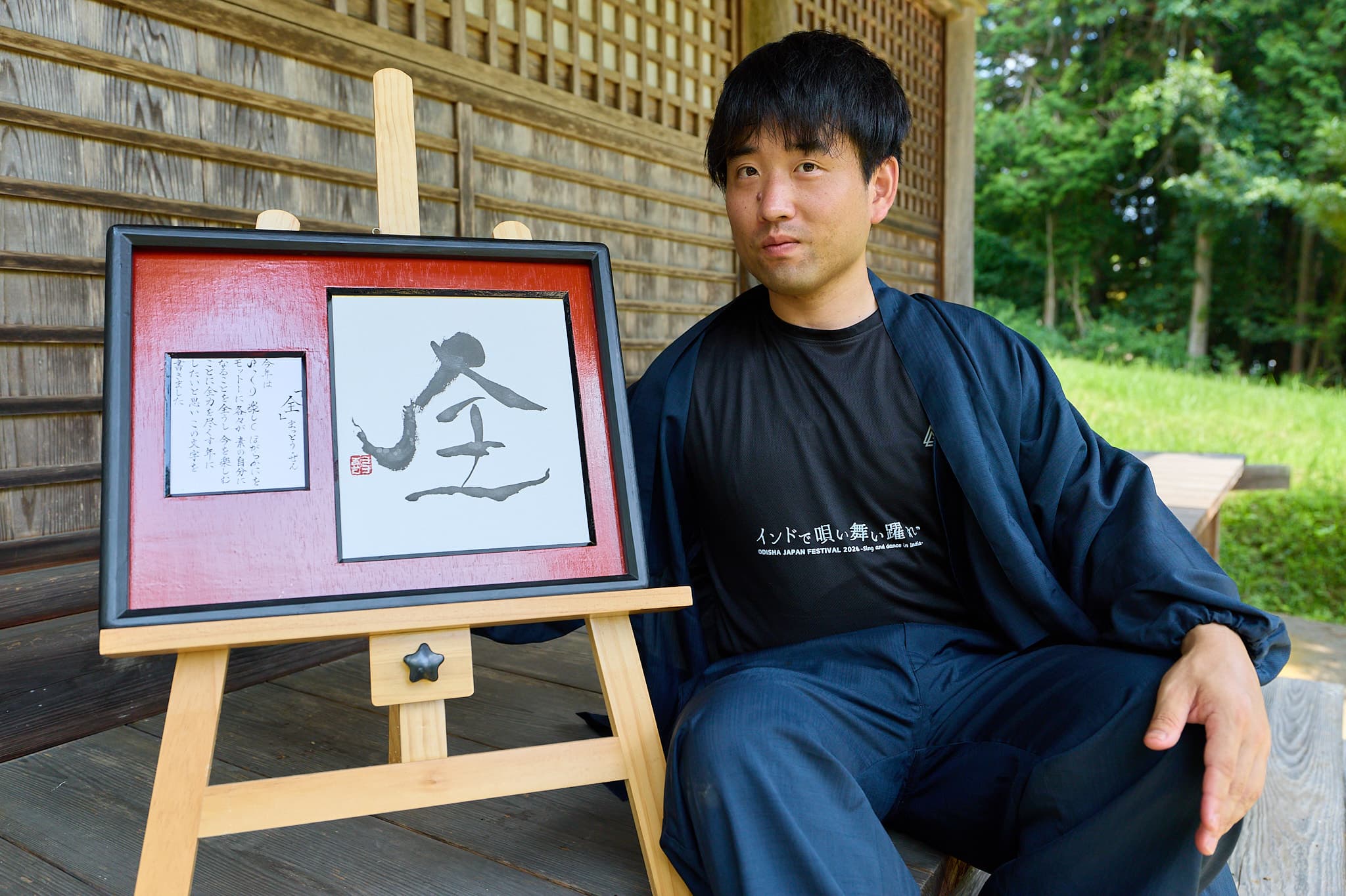 朱塗り額に収めた一文字「全」と解説書き。傍らに座る作務衣姿の森下友喜